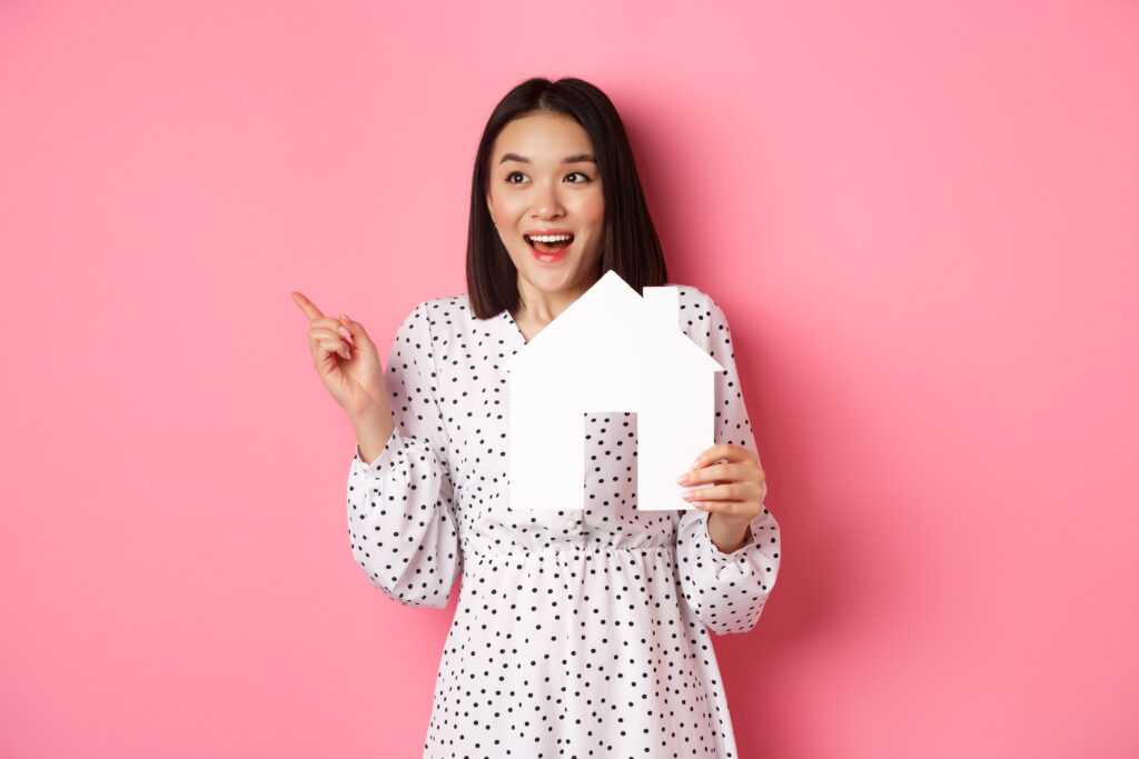 real-estate-excited-asian-woman-showing-paper-house-model-pointing-looking-left-copy-space-st