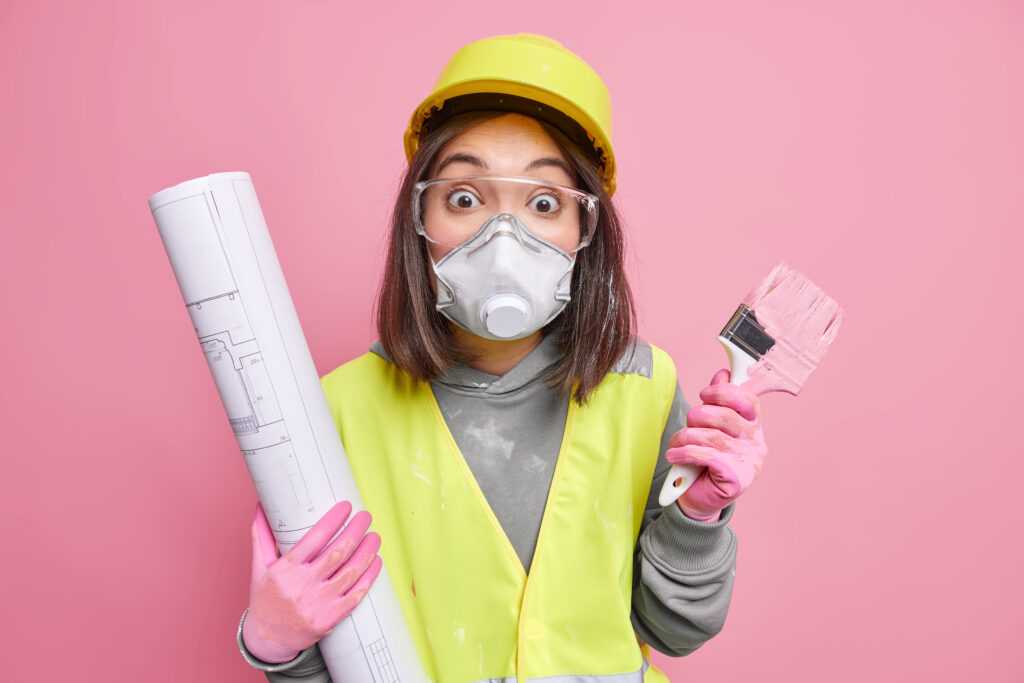 woman-wears-safety-glasses-respirator-hardhat-holds-blueprint-painting-brush-going-repaint-house-walls-poses-pink